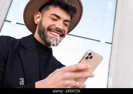 De dessous de jeune homme joyeux barbu dans le chapeau et l'habillement formel de navigation sur le téléphone cellulaire dans la ville Banque D'Images