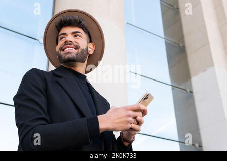 De dessous de jeune homme joyeux barbu dans le chapeau et l'habillement formel de navigation sur le téléphone cellulaire dans la ville Banque D'Images