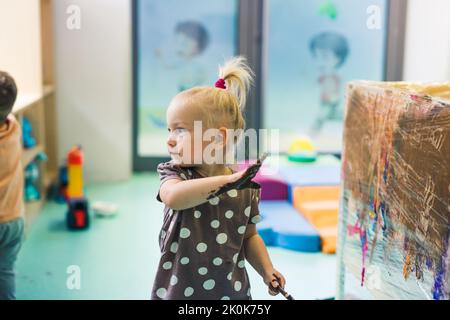 Peinture de film adhésif. Petite fille caucasienne enfant peinture avec ses bras sur un film adhésif enroulé tout autour de l'étagère en bois. Activité créative pour le développement des enfants à l'école maternelle. Photo de haute qualité Banque D'Images