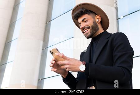 De dessous de jeune homme joyeux barbu dans le chapeau et l'habillement formel de navigation sur le téléphone cellulaire dans la ville Banque D'Images