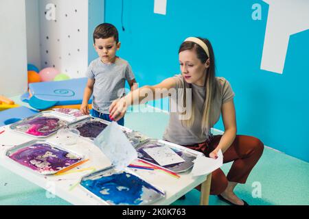 Jeu sensoriel à l'école maternelle multi-culturelle. Les tout-petits avec leur professeur jouant avec des pailles rayées et la peinture au lait, en utilisant la coloration de nourriture, le lait, le papier aquarelle, et les plateaux. Activité créative pour les enfants pour le développement du cerveau. Peinture au doigt. Photo de haute qualité Banque D'Images