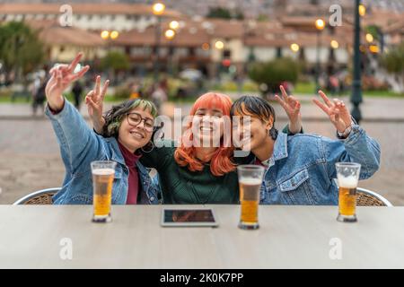 Jeunes amies gaies avec des verres de bière sur la table de repos dans le pub en plein air et montrant le geste de la victoire et regardant la caméra dans la ville de Cusco Banque D'Images