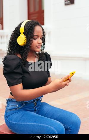 Femme ethnique gaie aux cheveux bouclés, écoutant de la musique dans un casque jaune et parcourant un smartphone dans la rue Banque D'Images