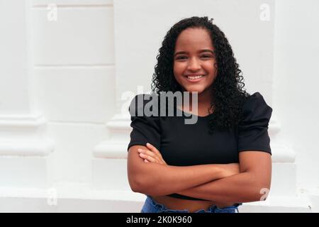 Jeune femme ethnique aux cheveux bouclés avec une tenue élégante, debout avec des bras croisés dans la rue de la ville et regardant l'appareil photo Banque D'Images