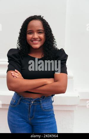 Jeune femme ethnique aux cheveux bouclés avec une tenue élégante, debout avec des bras croisés dans la rue de la ville et regardant l'appareil photo Banque D'Images