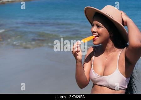 Jeune femme ethnique insouciante au bikini et au chapeau, se reposant sur une plage de sable, près de la mer en été Banque D'Images