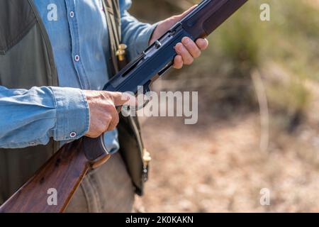 Crop mâle sans visage chasseur en camouflage gilet debout chargement fusil avec des balles en campagne sur fond flou Banque D'Images