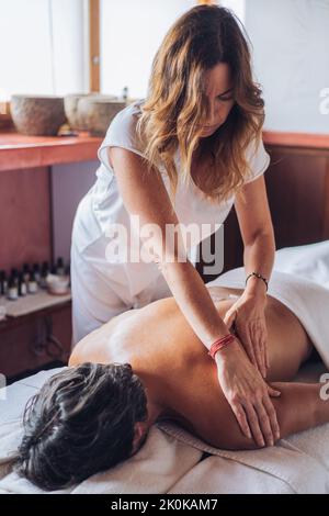Femme masseuse avec de longs cheveux en uniforme blanc faisant un massage du dos à l'homme allongé sur un canapé dans une salle de salon lumineuse pendant la journée Banque D'Images