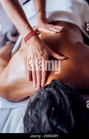 Cachez un masseur féminin non reconnaissable en uniforme blanc en faisant un massage du dos à un homme allongé sur un canapé dans une salle de salon lumineuse pendant la journée Banque D'Images