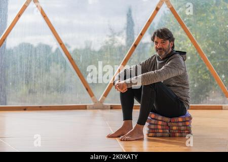 Vue latérale d'un homme gris à poils longs et joyeux, pieds nus et menton reposant sur une main, assis sur des couvertures pliées dans une pièce en bois, à proximité d'une vue panoramique Banque D'Images