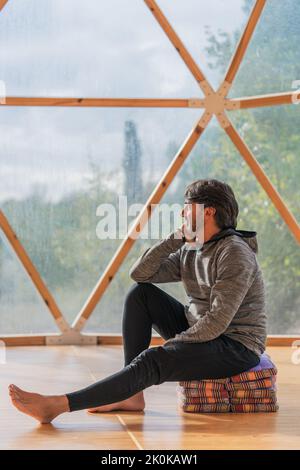 Vue latérale d'un homme gris à poils longs et joyeux, pieds nus et menton reposant sur une main, assis sur des couvertures pliées dans une pièce en bois, à proximité d'une vue panoramique Banque D'Images