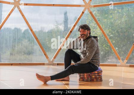 Vue latérale d'un homme gris à poils longs et joyeux, pieds nus et menton reposant sur une main, assis sur des couvertures pliées dans une pièce en bois, à proximité d'une vue panoramique Banque D'Images