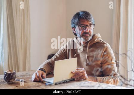 Vue latérale d'un homme à barbe d'un certain âge qui écrit dans un carnet tout en étant assis seul à la maison pendant la journée Banque D'Images