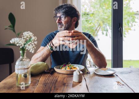 Un homme à cheveux gris barbu en verre, assis à une table en bois avec une assiette de vase avec des fleurs tenant une tasse de café et regardant un Banque D'Images