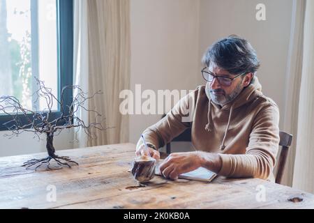 Vue latérale d'un homme à barbe d'un certain âge qui écrit dans un carnet tout en étant assis seul à la maison pendant la journée Banque D'Images