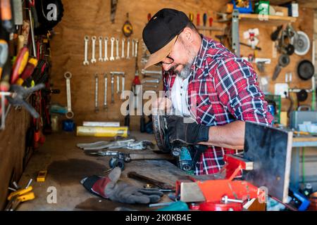 Vue latérale d'un homme d'âge moyen en casquette et chemise à carreaux, avec détail en bois sur une perceuse dans un garage léger Banque D'Images