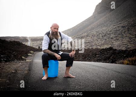 Homme avec costume est assis complètement désespéré et frappé dehors sur un bidon d'essence bleu quelque part dans le nulle part dans le désert volcanique à la fin o Banque D'Images