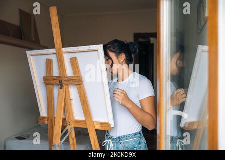 Femme ethnique en vêtements décontractés peint sur toile avec des peintures multicolores et pinceau dans une petite pièce légère Banque D'Images