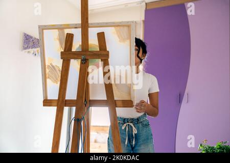 Femme ethnique en vêtements décontractés peint sur toile avec des peintures multicolores et pinceau dans une petite pièce légère Banque D'Images