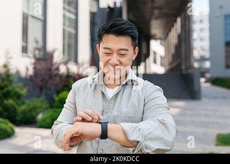 Homme asiatique mature et confiant regardant la montre, attendant le client près d'un immeuble de bureaux moderne, vérifiant le temps à l'extérieur Banque D'Images
