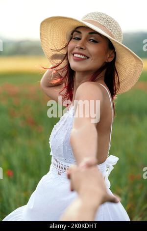 Vue latérale de la femme gaie dans la sundress et chapeau de paille tenant la récolte anonyme partenaire à la main tout en regardant l'appareil photo sur la prairie Banque D'Images