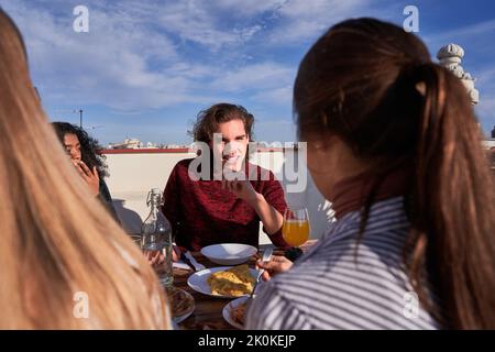 Positif jeunes amis divers portant des tenues décontractées en dégustant un délicieux déjeuner tout en se rassemblant à table sur la terrasse ensoleillée Banque D'Images