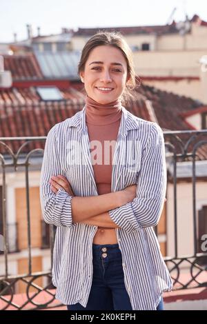 Jeune femme gaie dans une tenue décontractée debout avec des bras croisés sur la terrasse et regardant la caméra Banque D'Images