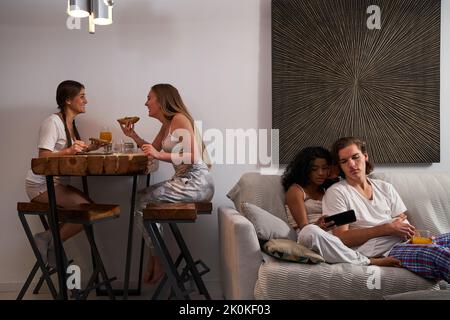 Un couple multiethnique souriant dans des vêtements domestiques qui naviguent sur un téléphone portable sur un canapé confortable près des femmes qui dînent dans le salon Banque D'Images