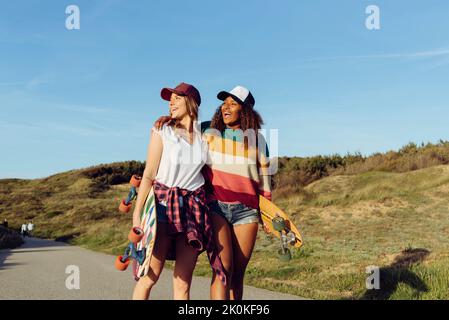 Deux belles patineuses multiethniques gaies tenant des planches de skate tout en marchant au milieu d'une route par une journée ensoleillée Banque D'Images