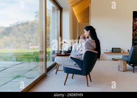 Vue latérale d'une jolie femme hispanique assise sur une chaise dans la chambre et regardant la grande fenêtre Banque D'Images