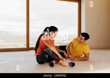 Gaie femme attrayante assise à côté d'un homme sportif reposant après avoir fait de l'exercice près d'une grande fenêtre dans la pièce Banque D'Images
