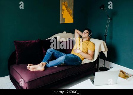 Homme caucasien attrayant couché avec les yeux fermés sur le canapé tout en ayant une sieste de musique sur les écouteurs à la maison Banque D'Images