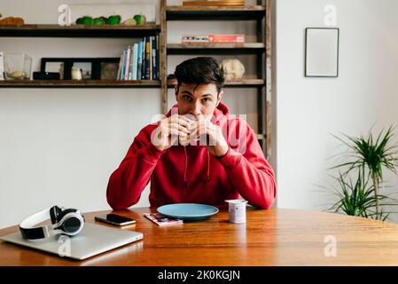 Beau homme caucasien heureux manger à la maison tout en ayant pause déjeuner de travailler avec l'ordinateur portable sur la table regardant l'appareil photo Banque D'Images
