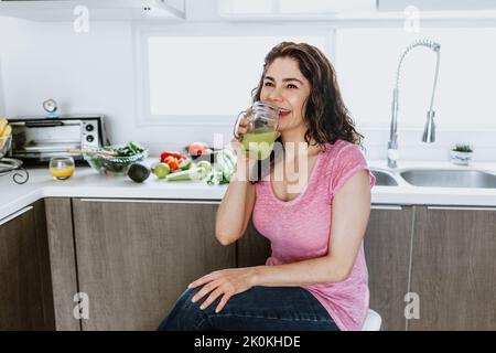 Positive femme avec des cheveux bouclés dans des vêtements décontractés assis dans la cuisine contre la table avec des légumes frais et boire un smoothie vert sain à la maison i Banque D'Images
