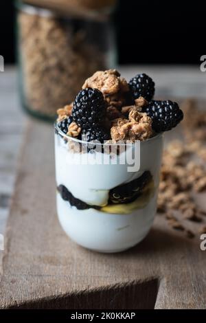 Vue en gros plan du verre avec granola de noix mélangée avec des bleuets et du yaourt placés sur une planche à découper en bois près du pot Banque D'Images