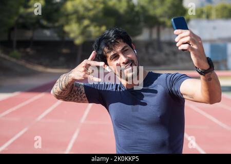 Joyeux coureur hispanique de vêtements de sport montrant le mouvement rock et se portant autoportrait sur un smartphone tout en se tenant au stade par beau temps Banque D'Images