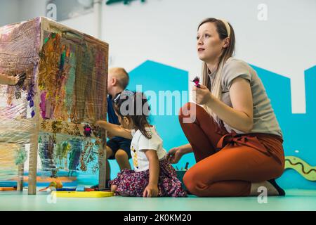 Peinture de film adhésif. Les tout-petits peignent avec une éponge, des pinceaux et des peintures sur un film adhésif enroulé autour de l'étagère en bois. Un enseignant les aidant. Activité créative pour les enfants développement des compétences sensorielles à la maternelle. Photo de haute qualité Banque D'Images