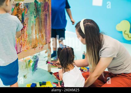 Peinture de film adhésif. Les tout-petits peignent avec une éponge, des pinceaux et des peintures sur un film adhésif enroulé autour de l'étagère en bois. Un enseignant les aidant. Activité créative pour les enfants développement des compétences sensorielles à la maternelle. Photo de haute qualité Banque D'Images