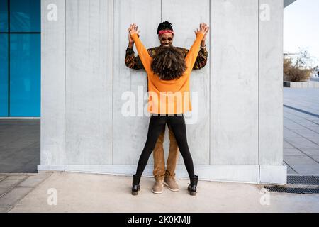 Vue arrière d'une femme afro-américaine élégante et méconnaissable avec des cheveux bouclés jouant et dansant avec l'homme dans des vêtements brillants contre un mur gris de rue Banque D'Images