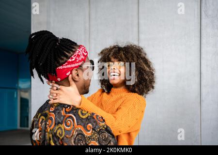 Vue arrière aimant Afro-américain moderne homme et femme embrassant et souriant avec des couleurs vives contre le mur gris dans la rue Banque D'Images