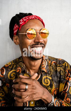 Joyeux Afro-américain avec des dreadlocks portant des lunettes de soleil rondes élégantes avec un bandeau rouge et une chemise colorée souriant loin Banque D'Images