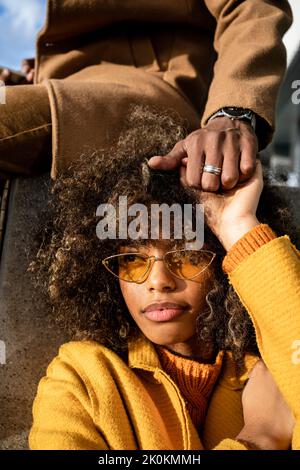 Jeune femme ethnique élégante avec des boucles afro portant des lunettes de soleil tendance et un manteau jaune tenant la main de crop boyfriend à la lumière du soleil Banque D'Images