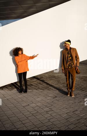 Un beau couple africain branché debout dans la rue portant des lunettes de soleil penchées sur un mur blanc regardant l'un l'autre dans la lumière du coucher du soleil Banque D'Images