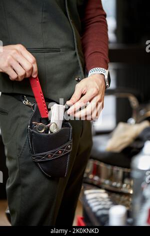 Vue latérale d'un petit barbier masculin méconnaissable avec divers instruments professionnels debout dans un salon de coiffure Banque D'Images