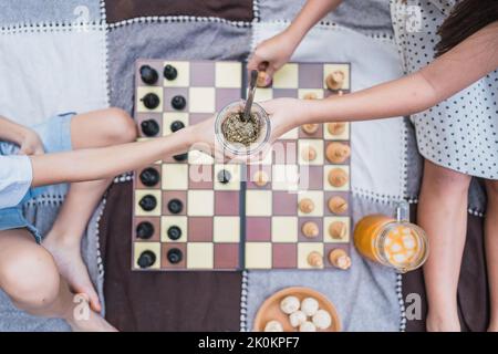 Vue de dessus de la récolte anonyme garçon recevant du thé à base de plantes de la sœur sur des pièces d'échecs sur le tissu pendant le pique-nique Banque D'Images