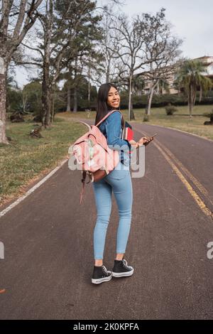 Vue arrière du corps d'une jeune étudiante de l'ethnie heureuse avec sac à dos et livres à l'aide d'un téléphone portable et regardant l'appareil photo au-dessus de l'épaule tout en se promenant Banque D'Images
