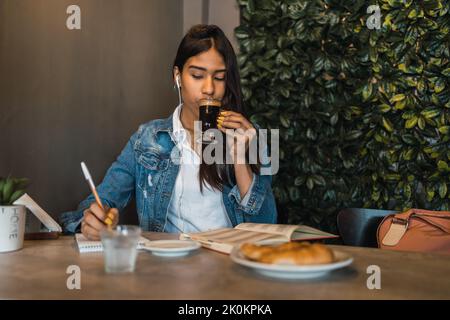 Contenu intelligent jeune hispanique étudiante écrire dans le livre d'auteur tout en faisant devoirs à la table boire du café Banque D'Images