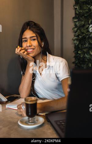 Femme hispanique gaie avec écouteurs étudiant en ligne et regardant la vidéo conférence sur ordinateur portable et prenant des notes dans le livre d'auteur à la table avec une tasse de café Banque D'Images