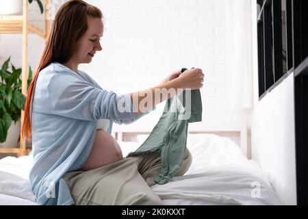 Mère enceinte tenant des vêtements de bébé dans les mains, bonne femme enceinte attendant l'enfant. Préparation à l'accouchement, fille grand ventre avancé grossesse Banque D'Images