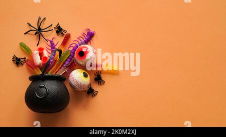 Gros plan de bonbons gélifiés, araignées, yeux, mâchoires, vers et chauves-souris dans un pot d'Halloween sur fond orange, vue de dessus, plat, espace de copie. Décor conce Banque D'Images